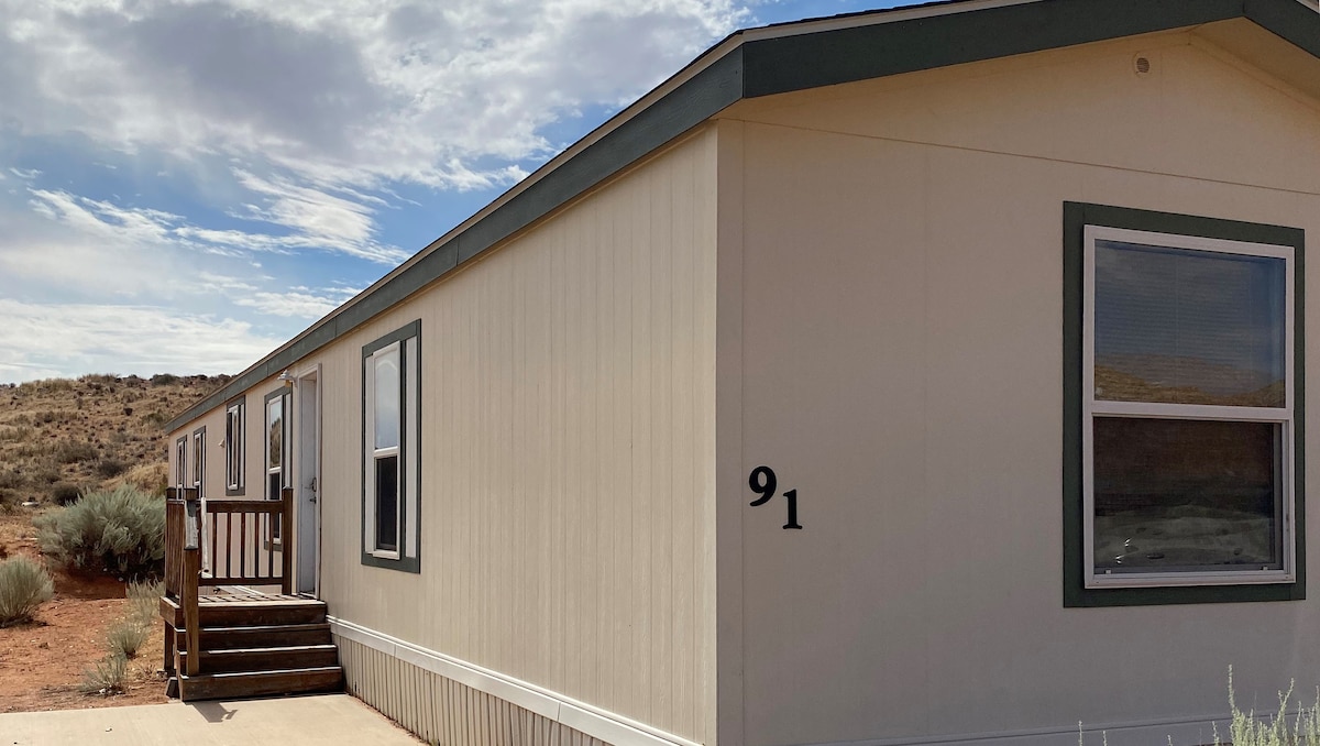 A single-story building is shown, featuring a light exterior with contrasting trim. Steps lead up to a welcoming entrance. Two windows are visible, allowing natural light to enhance the interior. The surrounding landscape showcases a desert terrain, providing a serene backdrop.