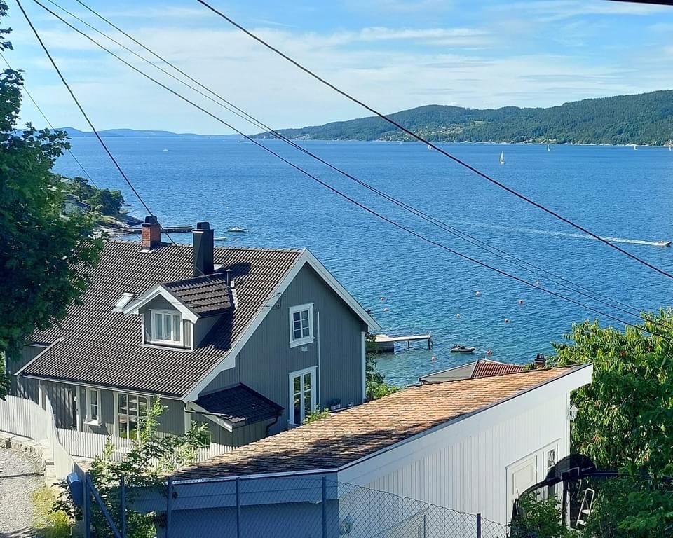A view of the Oslofjord is captured, showcasing calm waters and distant hills under a clear blue sky. The property is nestled nearby, featuring traditional architecture with a sloped roof. Nearby boats can be seen along the shoreline, enhancing the serene coastal environment.