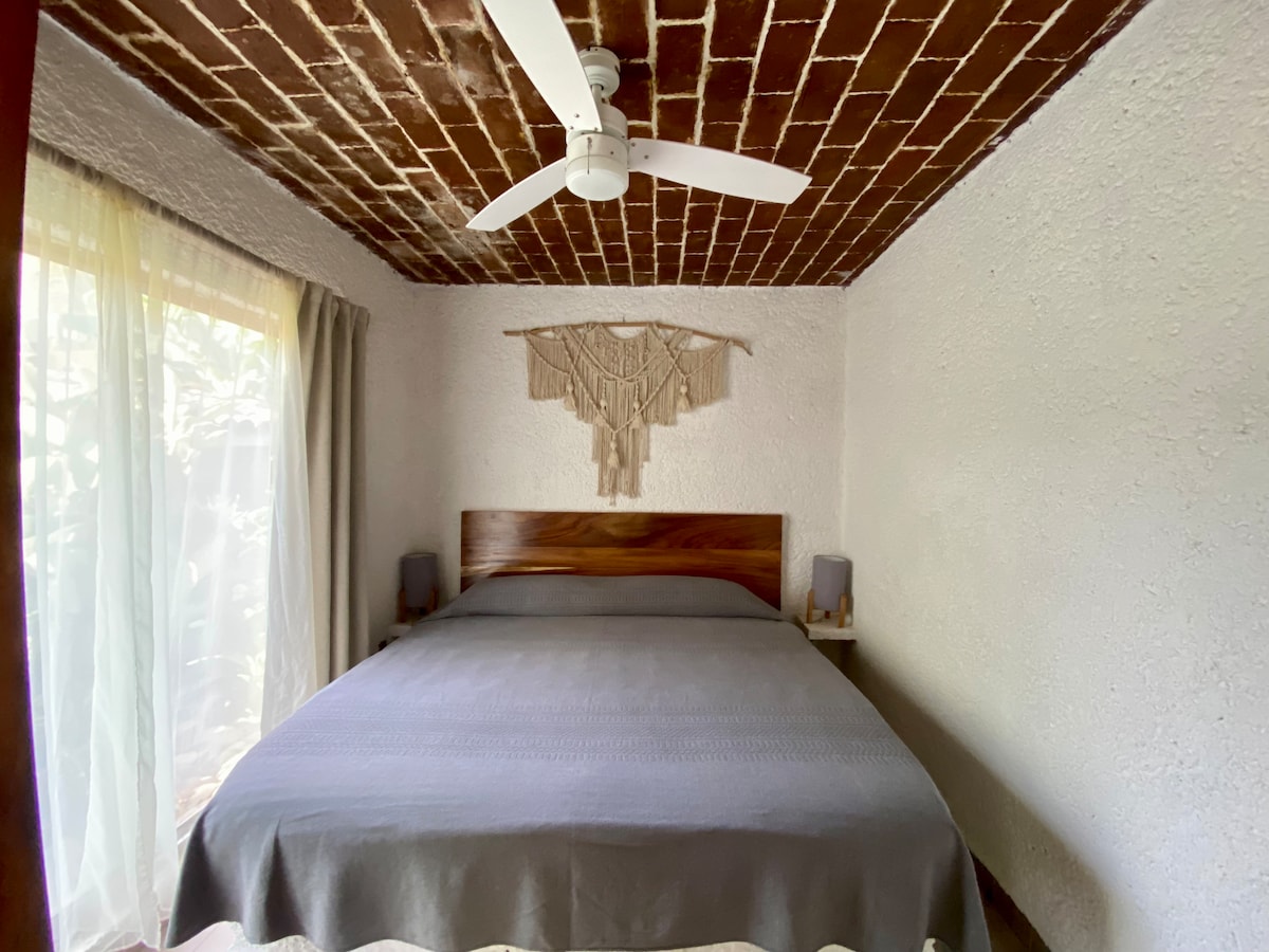 A spacious sleeping area features a large bed with a light grey blanket and decorative wall art. Natural light enters through a large window partly covered by curtains, while a ceiling fan ensures comfort.