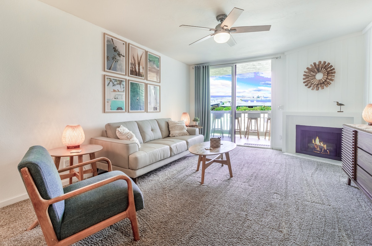 A modern coastal living area is illuminated by natural light from sliding glass doors, opening to a balcony. A comfortable gray sofa is centered in the space, accompanied by a wooden coffee table and stylish armchair. Artful wall decor complements the serene atmosphere.