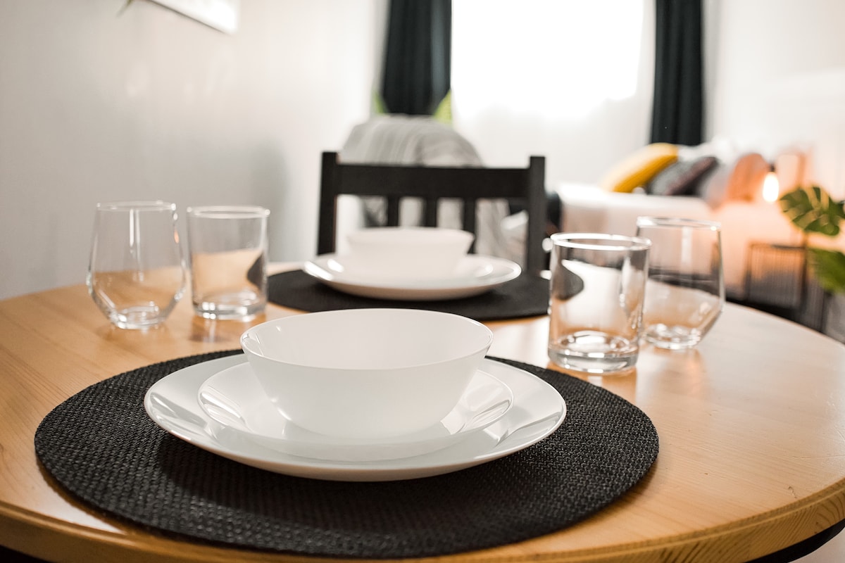 A dining table is set with white plates and bowls, accompanied by glassware. A dark woven placemat is beneath the dishes. The background features a softly lit living area with a cozy sofa and light curtains, enhancing the welcoming ambiance.