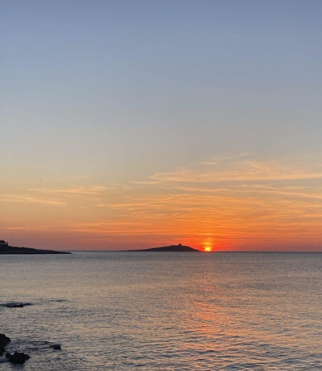 A serene seascape is captured at sunset, with the sun dipping below the horizon, casting warm hues across the sky and reflecting on the calm water. An island silhouette is visible in the distance, enhancing the tranquil atmosphere.