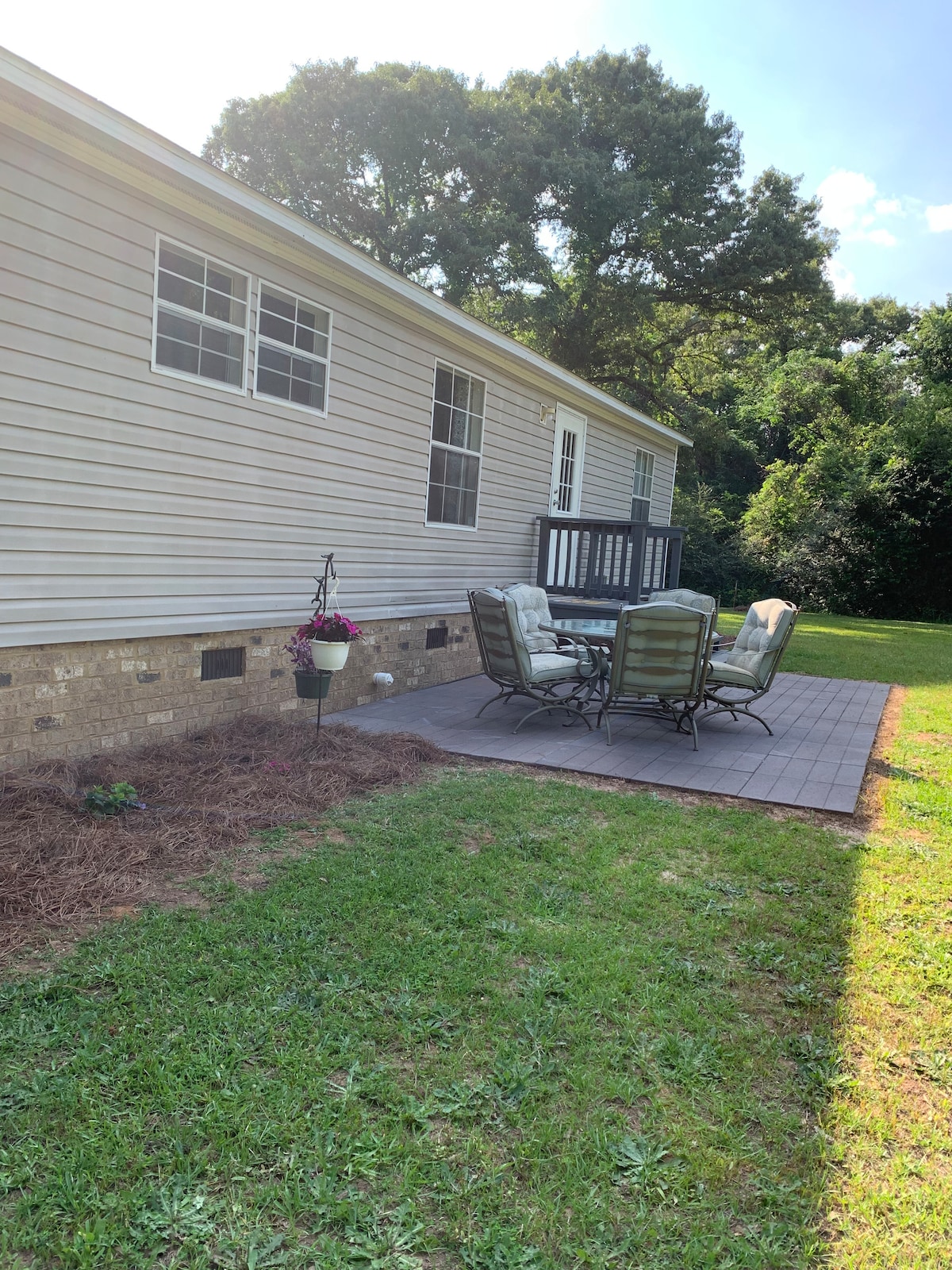 An outdoor patio area is shown, featuring a table with four cushioned chairs. A hanging planter with flowers adds a touch of color. The patio is surrounded by a grassy yard, leading to a wooded area in the background.