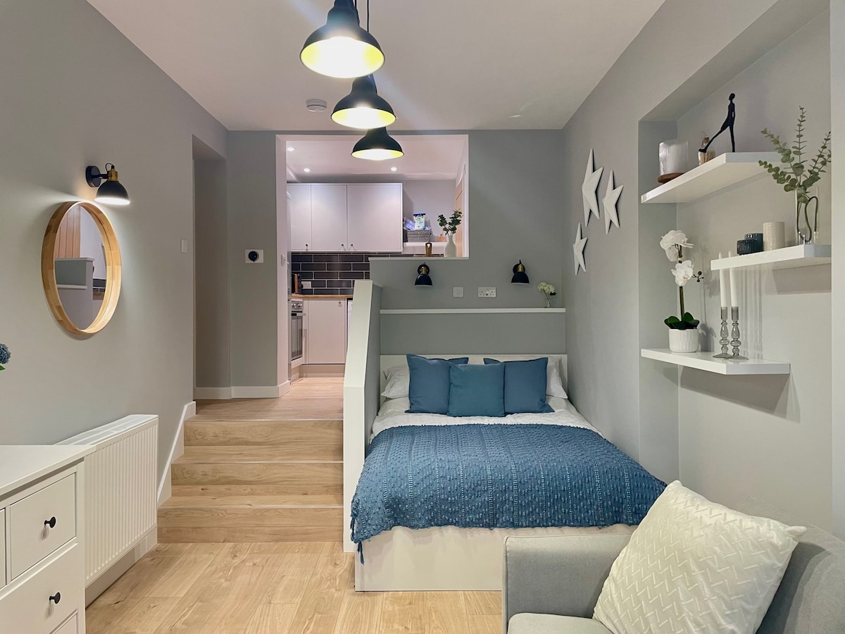 An open-plan living space features a comfortable settee and a cozy sleeping area. A decorative blue blanket is laid over the spacious bed, which is accentuated by soft blue pillows. Light fixtures hang from the ceiling, illuminating the room with a warm glow.