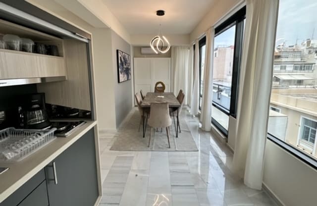 A modern dining area features a wooden table surrounded by six upholstered chairs. Soft, natural light filters through large windows, illuminating the marble floor. The kitchenette with sleek cabinetry provides a functional space for meal preparation.