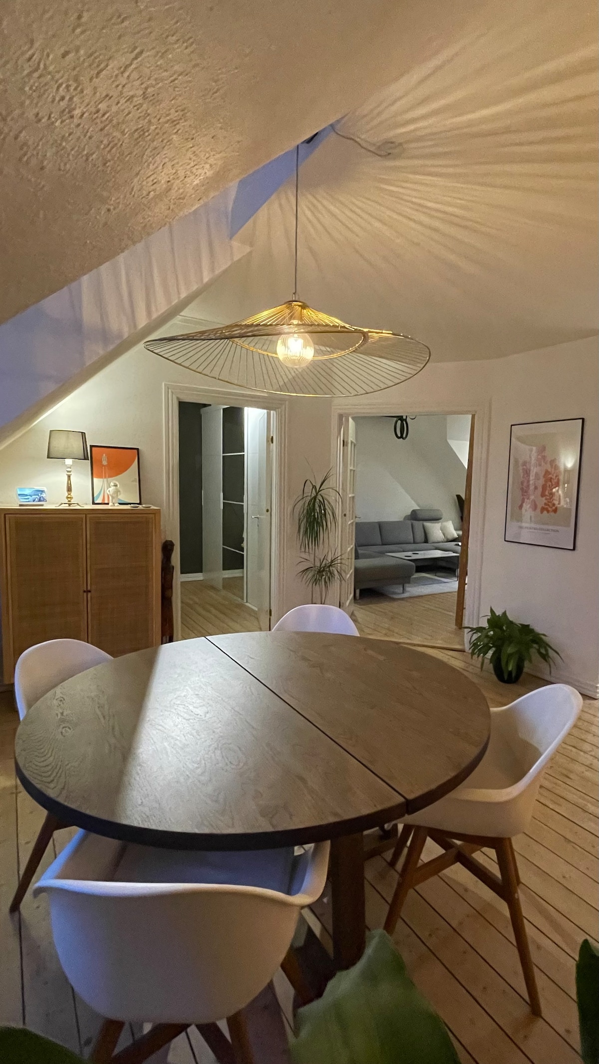 A circular dining table is surrounded by four white chairs, situated on wooden flooring. A woven pendant light hangs above, casting soft shadows on the ceiling. Green plants are positioned nearby, with a glimpse of a doorway leading to another room in the background.