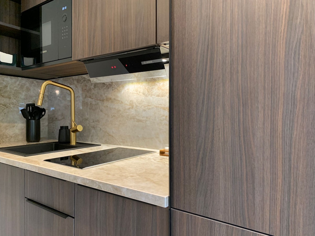 The kitchen features sleek wooden cabinets and a marble countertop, complemented by modern appliances. A stylish faucet in gold accents the space, while subtle under-cabinet lighting enhances visibility. Cooking utensils are neatly arranged in a container, adding to the functional design.