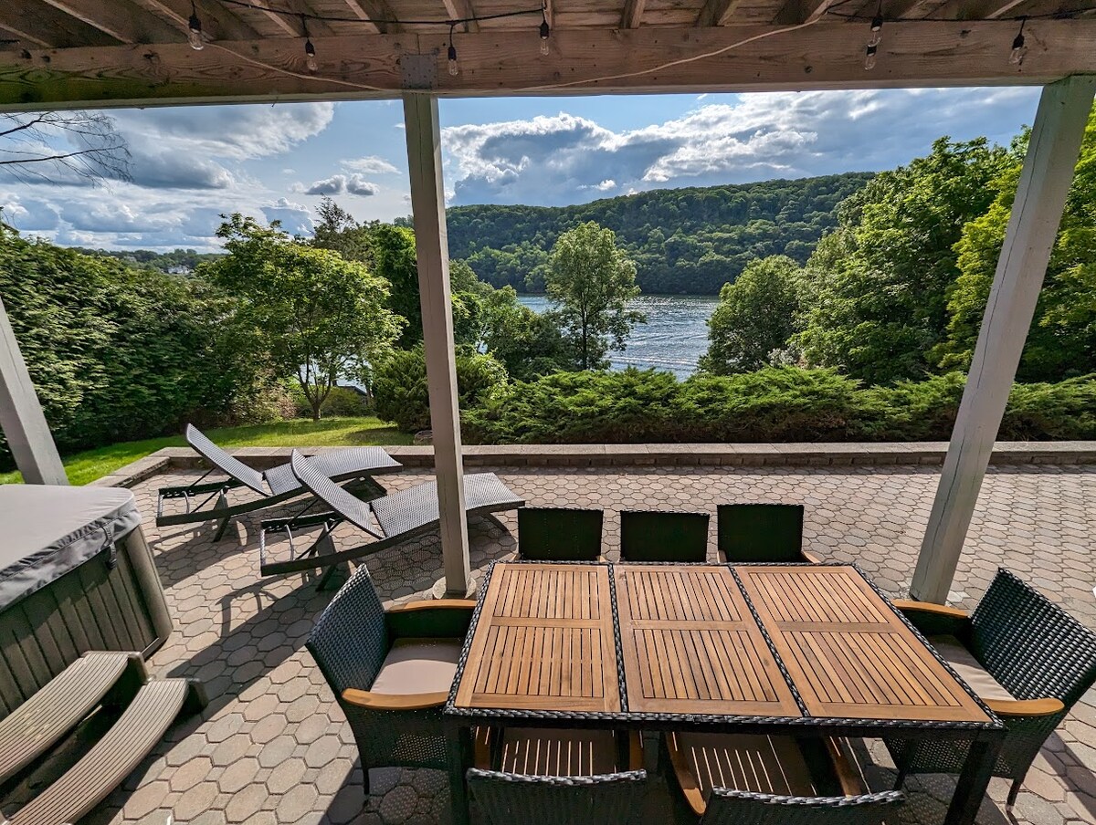 An outdoor patio area features a wooden dining table surrounded by several chairs, complementing a hot tub nearby. Comfortable lounge chairs are positioned for lake views, enhancing the serene natural setting framed by trees and hills under a partly cloudy sky.