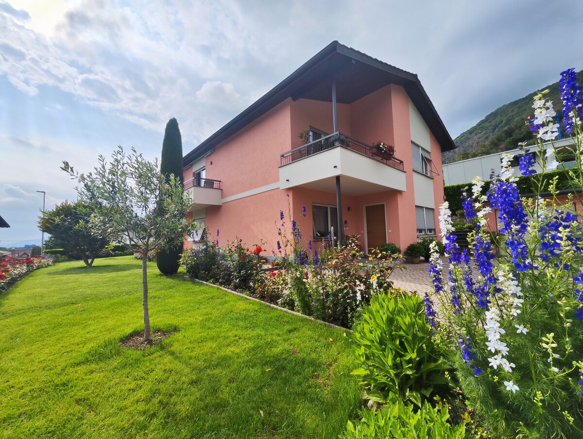 A two-story building is showcased, featuring a light pink exterior and multiple balconies adorned with potted plants. The surrounding garden is lush with vibrant flowers, including tall blue and white blooms, set against a backdrop of green grass and neatly landscaped shrubs.