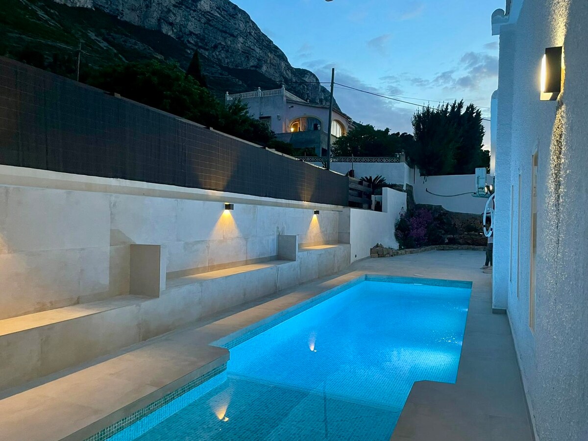 The pool area is illuminated with soft lighting, highlighting a clear blue pool surrounded by smooth stone surfaces. A dramatic mountain backdrop is visible, adding a sense of serenity. Built-in seating is situated along one side of the pool, enhancing relaxation options.