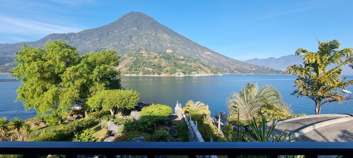 A panoramic view showcases a tranquil lake framed by lush greenery and mountains. Multiple plants and trees populate the foreground, leading to the water's edge. The clear sky and distant peaks enhance the serene atmosphere of the setting.