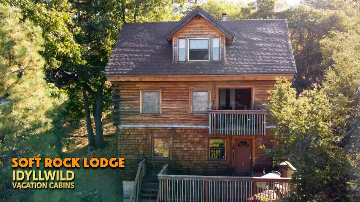 The cabin, constructed of natural wood, is surrounded by lush greenery. A spacious front deck extends from the entrance, while multiple windows reflect a welcoming ambiance. The roof features a classic A-frame structure, enhancing the rustic charm of this inviting retreat.