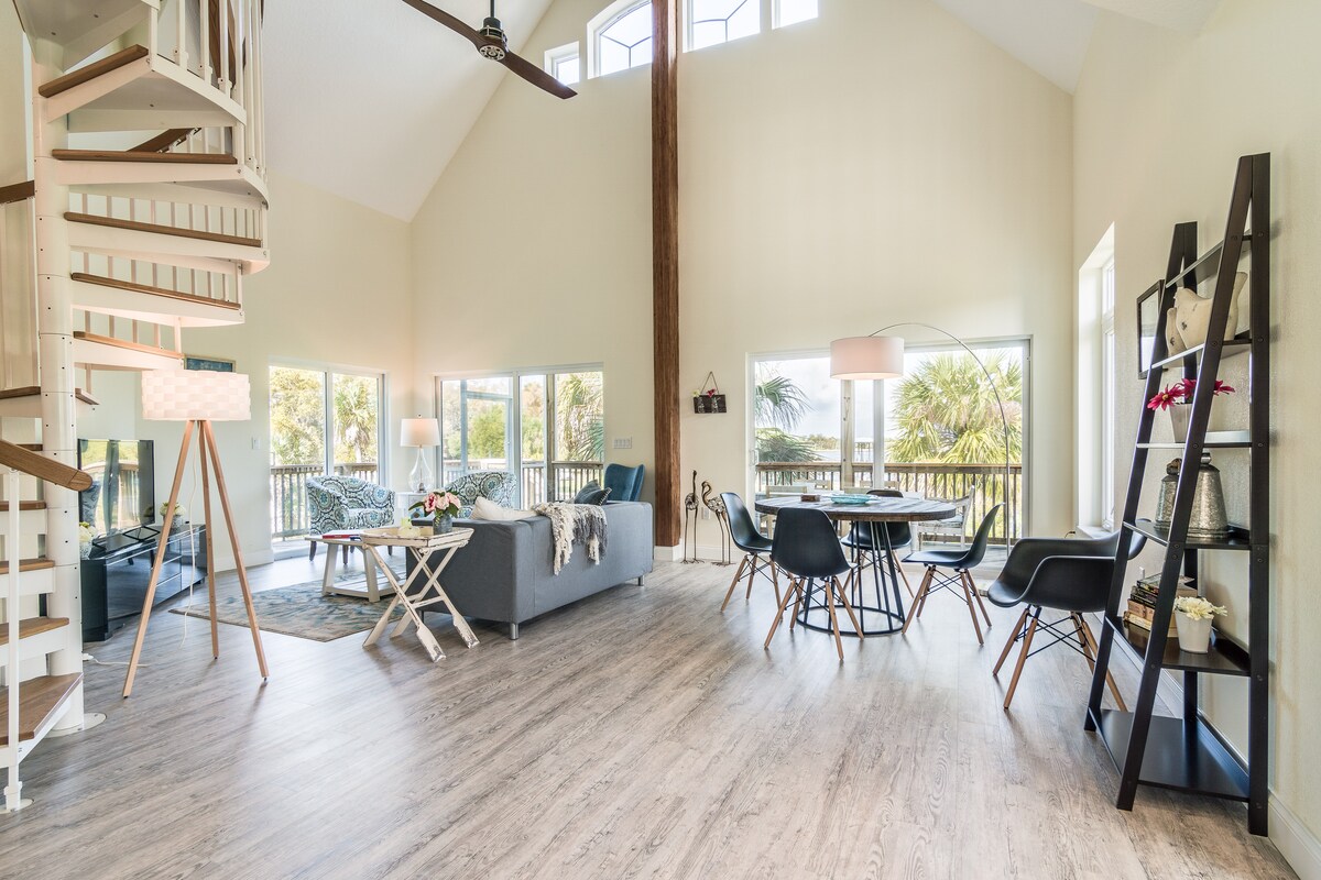An open living space showcases high ceilings and large windows that flood the room with natural light. A spiral staircase leads to the second floor. Modern furniture is arranged around a table and cozy seating area, fostering a welcoming environment with a view of the surrounding greenery.