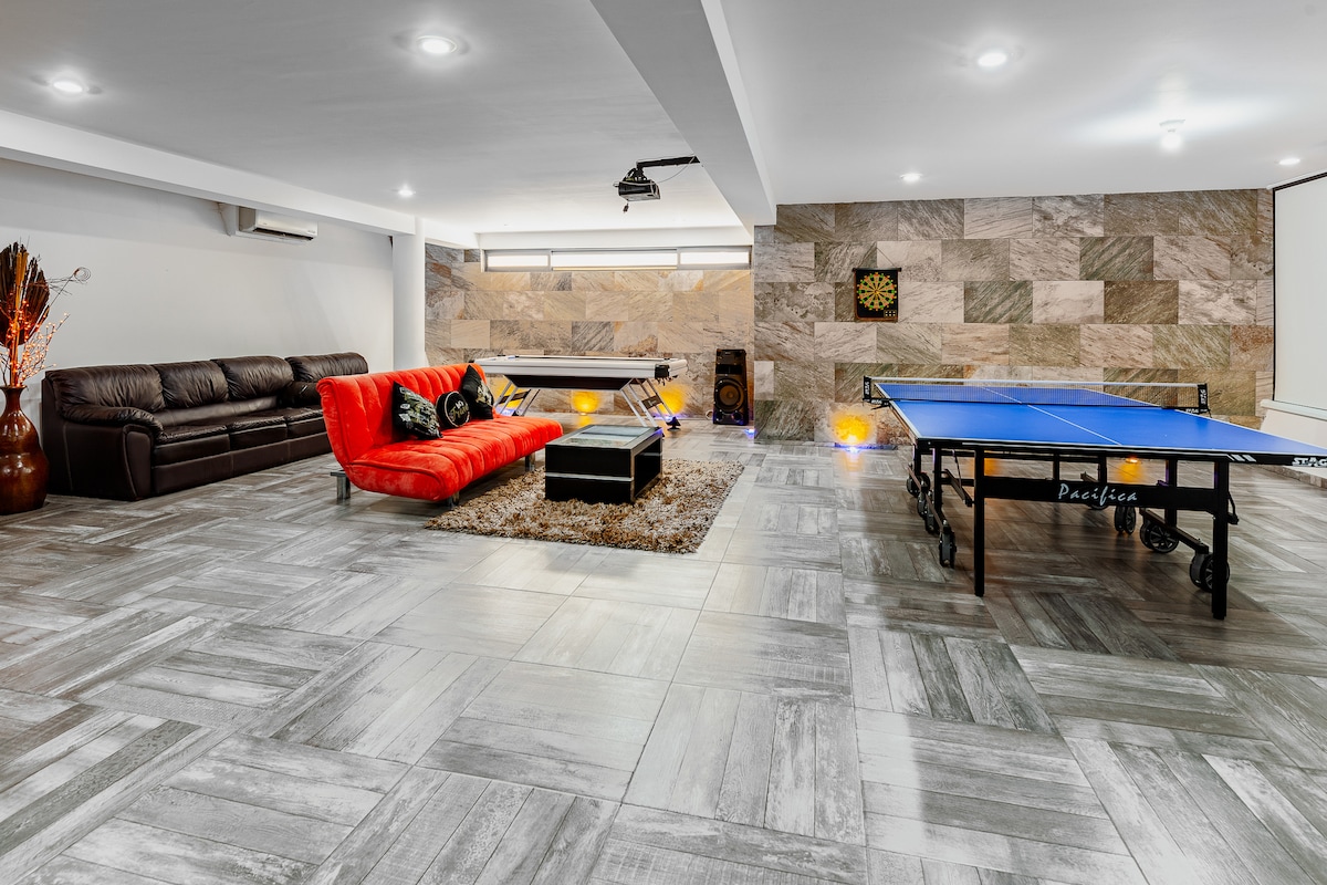 A game room is highlighted by a vibrant red sofa and a sleek wooden coffee table, positioned on a plush area rug. A ping pong table occupies one side, while a wall of textured stone adds visual interest. Soft overhead lighting illuminates the recreational space.