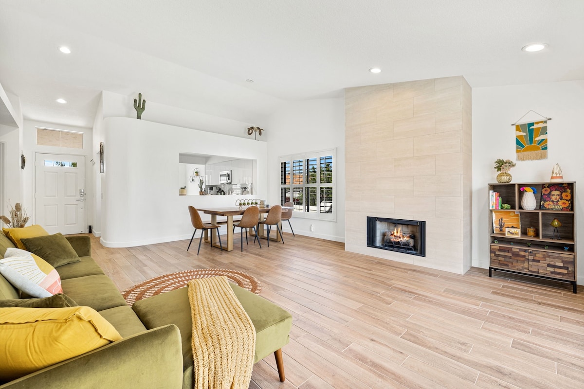 A spacious living area features a green sofa paired with brown dining chairs, situated near a modern fireplace. Large windows provide natural light, while tasteful decor elements, including a wall art piece and potted plants, enhance the inviting ambiance.