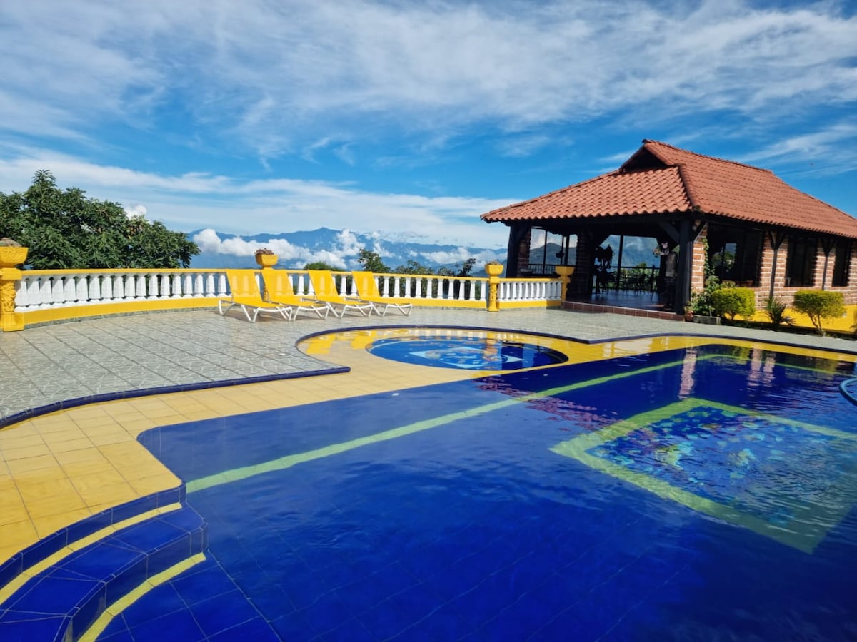 A large outdoor pool features vibrant blue tiles and is complemented by a separate circular jacuzzi. Sun loungers are positioned along the poolside, and a gazebo with a terracotta roof provides shaded seating. Lush greenery surrounds the area, with distant mountains visible under a partly cloudy sky.