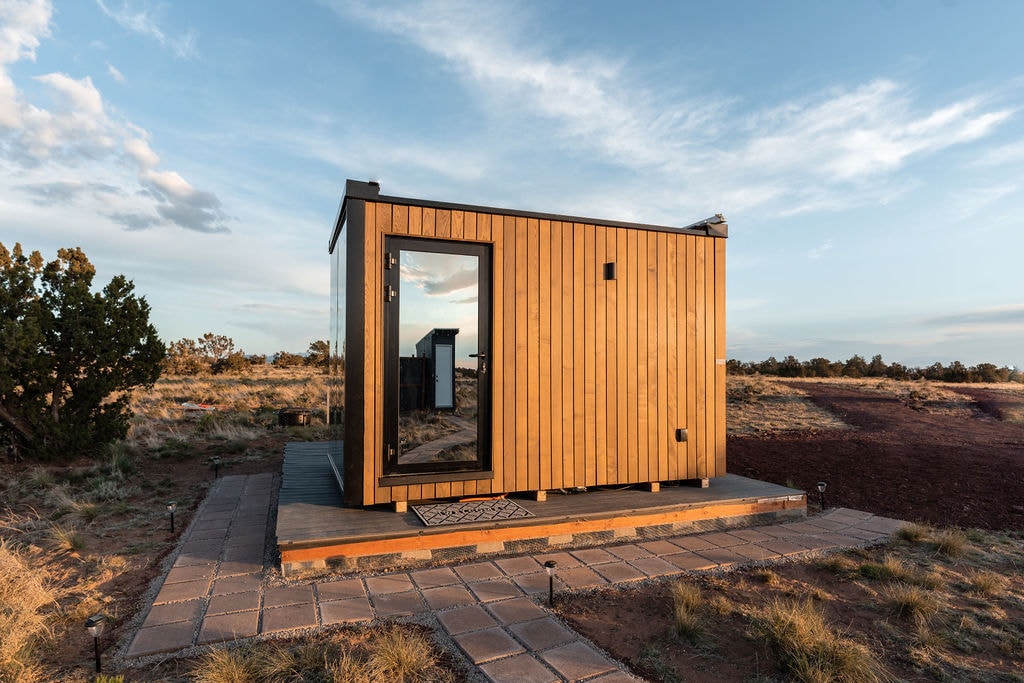 A tiny home with natural wood siding is set on a stone patio, blending into the landscape. Large windows reflect the surrounding environment, providing views of the open sky. The entry door is framed in black, adding a modern touch to its rustic charm.