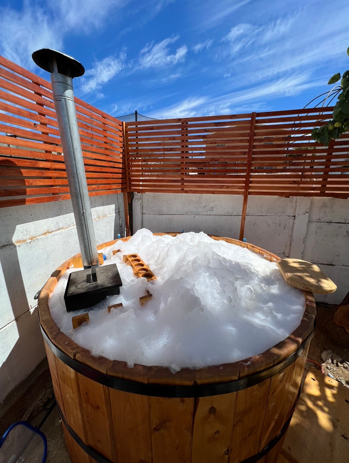 A wooden hot tub is filled with bubbles and surrounded by a wooden slatted privacy screen. A metal chimney extends from the tub, contributing to a peaceful outdoor setting under a blue sky.