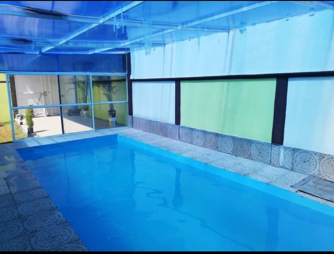 A swimming pool is enclosed by glass walls, allowing natural light to fill the space. The pool is surrounded by neatly tiled flooring, contributing to a serene atmosphere. A subdued color palette of blues and greens enhances the calming environment.