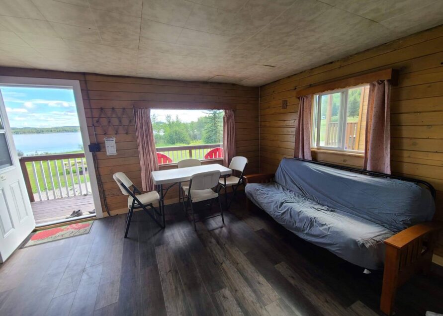 An inviting interior space is displayed, featuring a futon in the corner and a dining table with chairs positioned near a large window. Natural light enters the room, highlighting the wooden walls. Outdoors, a patio with red chairs can be seen, alongside a scenic view of the lake.