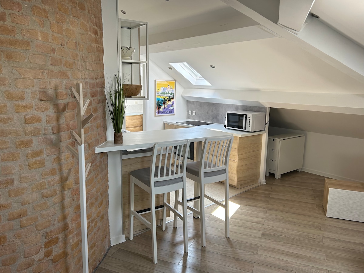 An open kitchen area features light wooden cabinetry and a microwave. A white bar table with two gray stools sits against a rustic brick wall, offering a casual dining space. Natural light streams in through a skylight, enhancing the airy atmosphere.