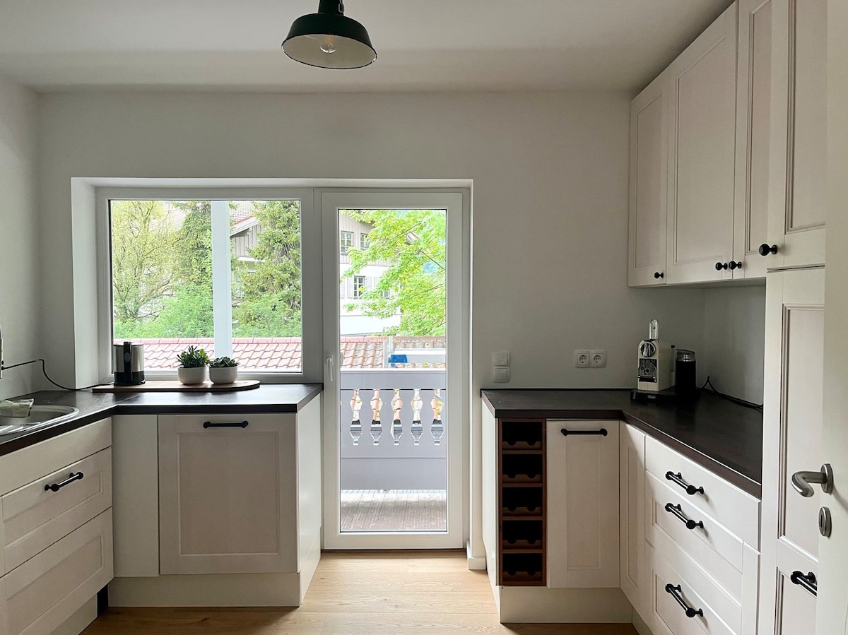 A modern kitchen is presented, featuring white cabinetry with sleek black hardware and ample counter space. Large windows provide natural light and a view of greenery outside, while a wine rack and various kitchen appliances are visible on the counters.