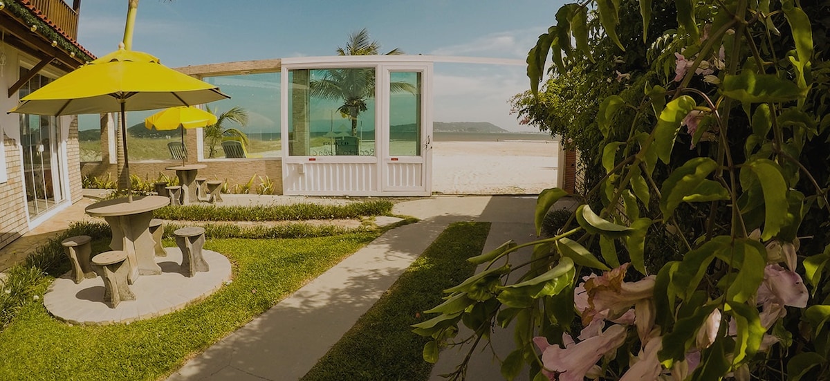 A garden area features a round stone table with matching benches under a yellow umbrella. Lush greenery and flowering plants frame the space. Beyond, a large glass wall offers a view of the beach and distant water, enhancing the connection between indoor and outdoor living.