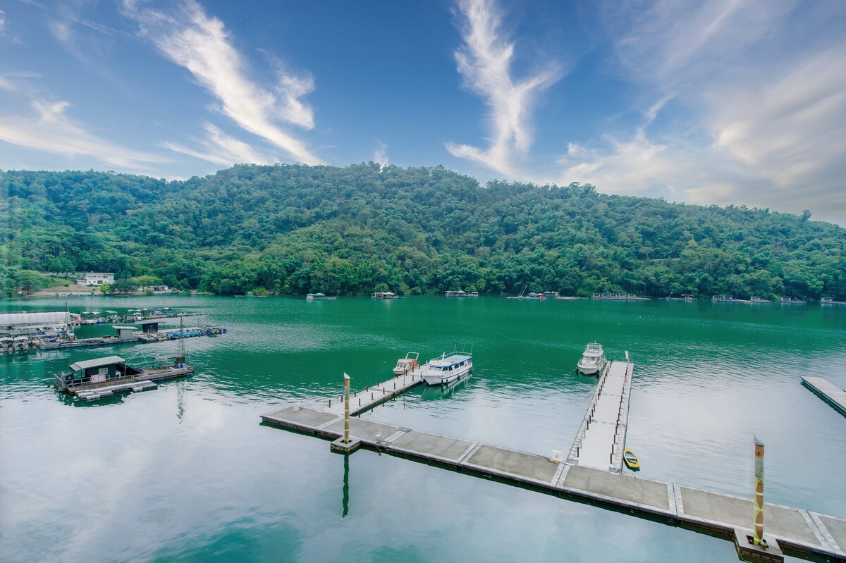 A serene view of a marina situated on a tranquil lake, surrounded by lush green hills. The calm water reflects the sky and boats are docked along the piers, creating a peaceful atmosphere.