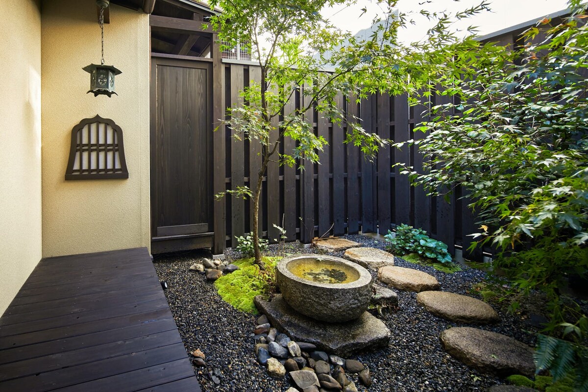 A serene outdoor space features a gravel pathway leading to a stone basin filled with water. Nearby, rounded stepping stones provide a natural transition through the garden, surrounded by lush greenery. A wooden fence offers privacy, while a lantern hangs gracefully from the wall.
