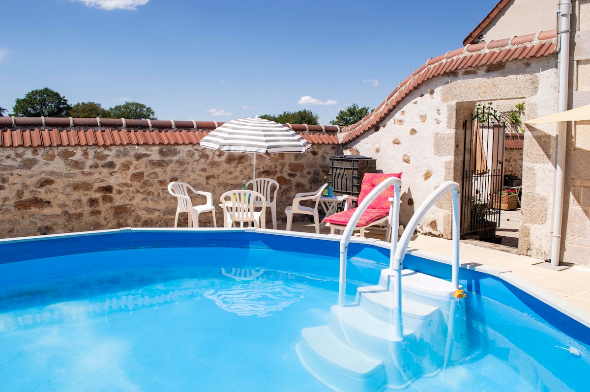 An oval pool with a depth of 1.25m is situated in a sunlit yard. Surrounding the pool are several white lounge chairs and a striped umbrella, providing shaded seating. A stone wall and an entrance gate feature in the background, enhancing the tranquil setting.