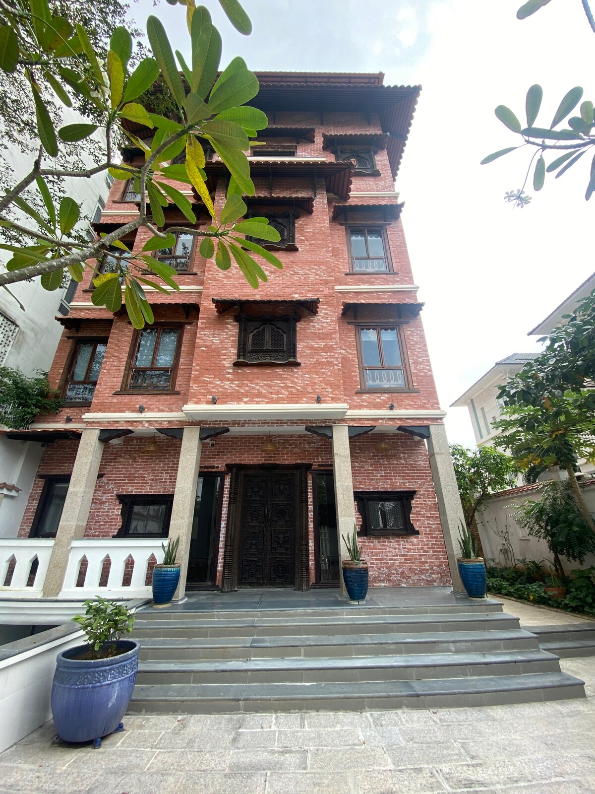 The exterior of the recently constructed house features a warm red brick facade. A multi-story structure is framed by large windows and traditional wooden elements. Green plants are placed along the entrance, highlighting the inviting staircase leading to the front door.
