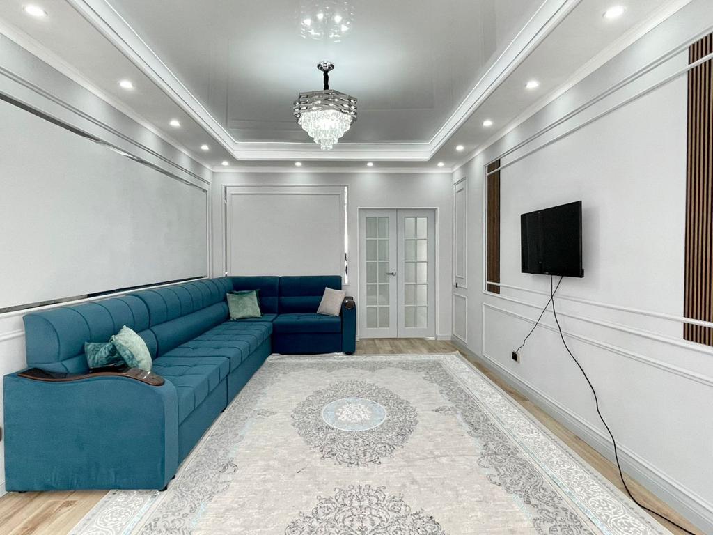 The spacious living room features a large blue sectional sofa positioned against a light wall. A stylish chandelier hangs from the ceiling, providing soft illumination. A flat-screen television is mounted on the wall, and a decorative area rug adds a touch of elegance to the hardwood floor.