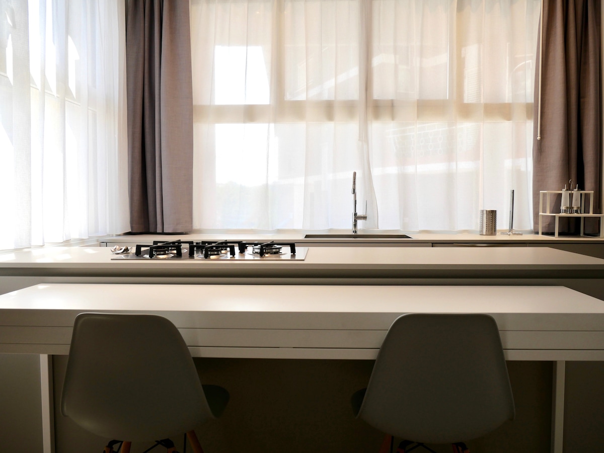 A well-lit modern kitchen area features a long white countertop with a built-in gas stove. Two minimalist chairs are positioned at the edge of the counter. Sheer curtains filter natural light, offering a bright and airy atmosphere.