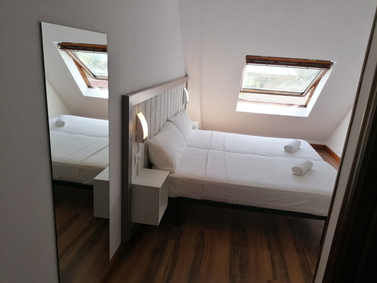 A well-lit room features a spacious bed with crisp white linens and two towels neatly arranged. Natural light filters in through two slanted windows, illuminating the warm wood flooring. A mirrored accent reflects the inviting atmosphere of the room, enhancing the sense of space.