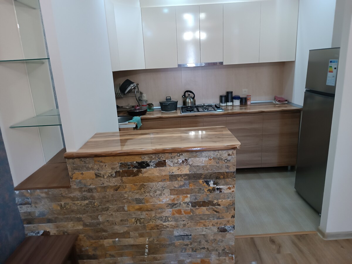 A modern kitchen features sleek cabinetry and a wooden countertop. Cooking appliances are positioned near the wall, complemented by a silver refrigerator. Shelves display various utensils, while natural light brightens the space.