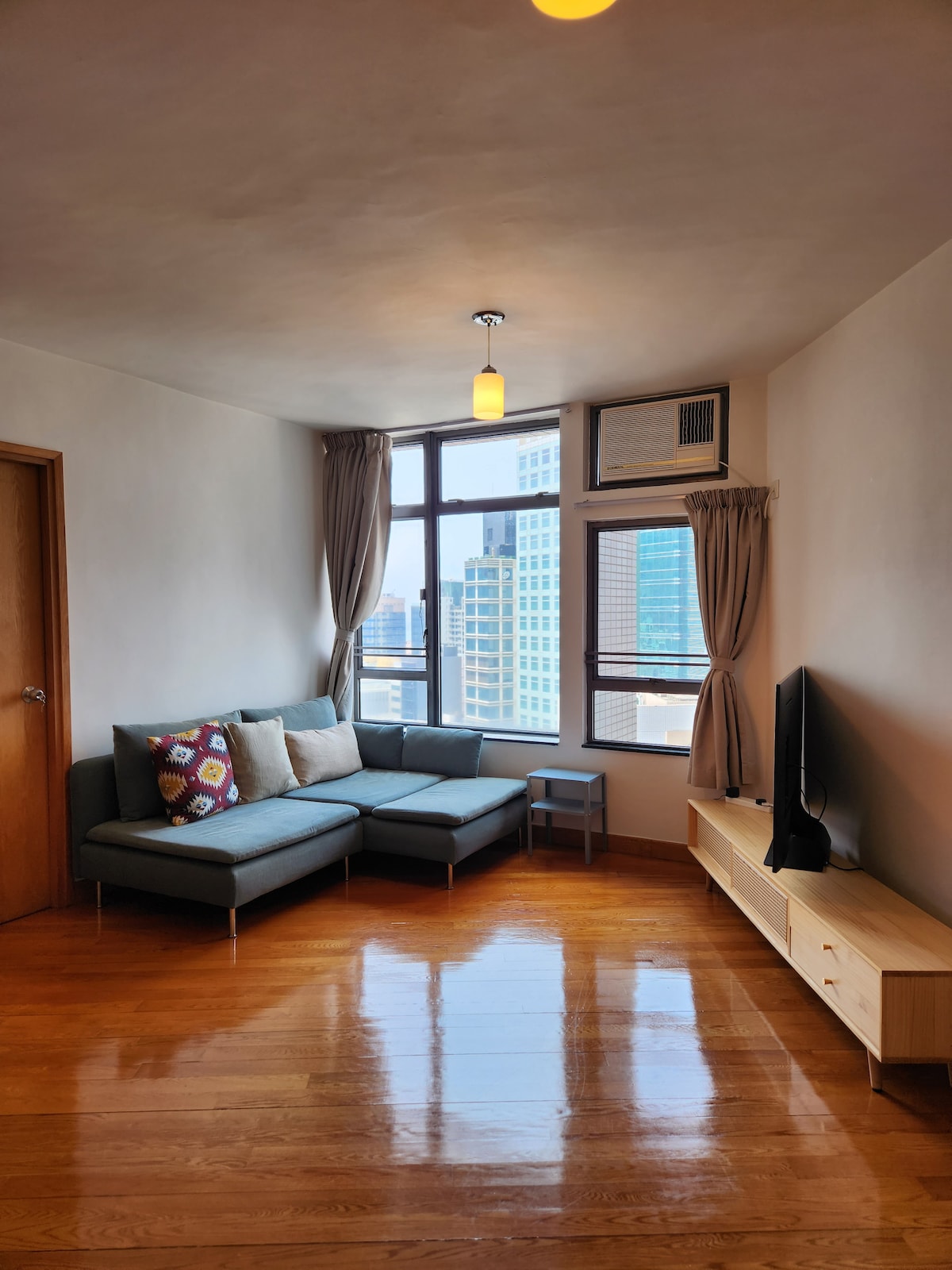 A comfortably arranged living area features a modern sectional sofa adorned with decorative pillows. A flat-screen television rests on a light wooden console, while large windows allow ample natural light to fill the space, highlighting the polished wooden floor.