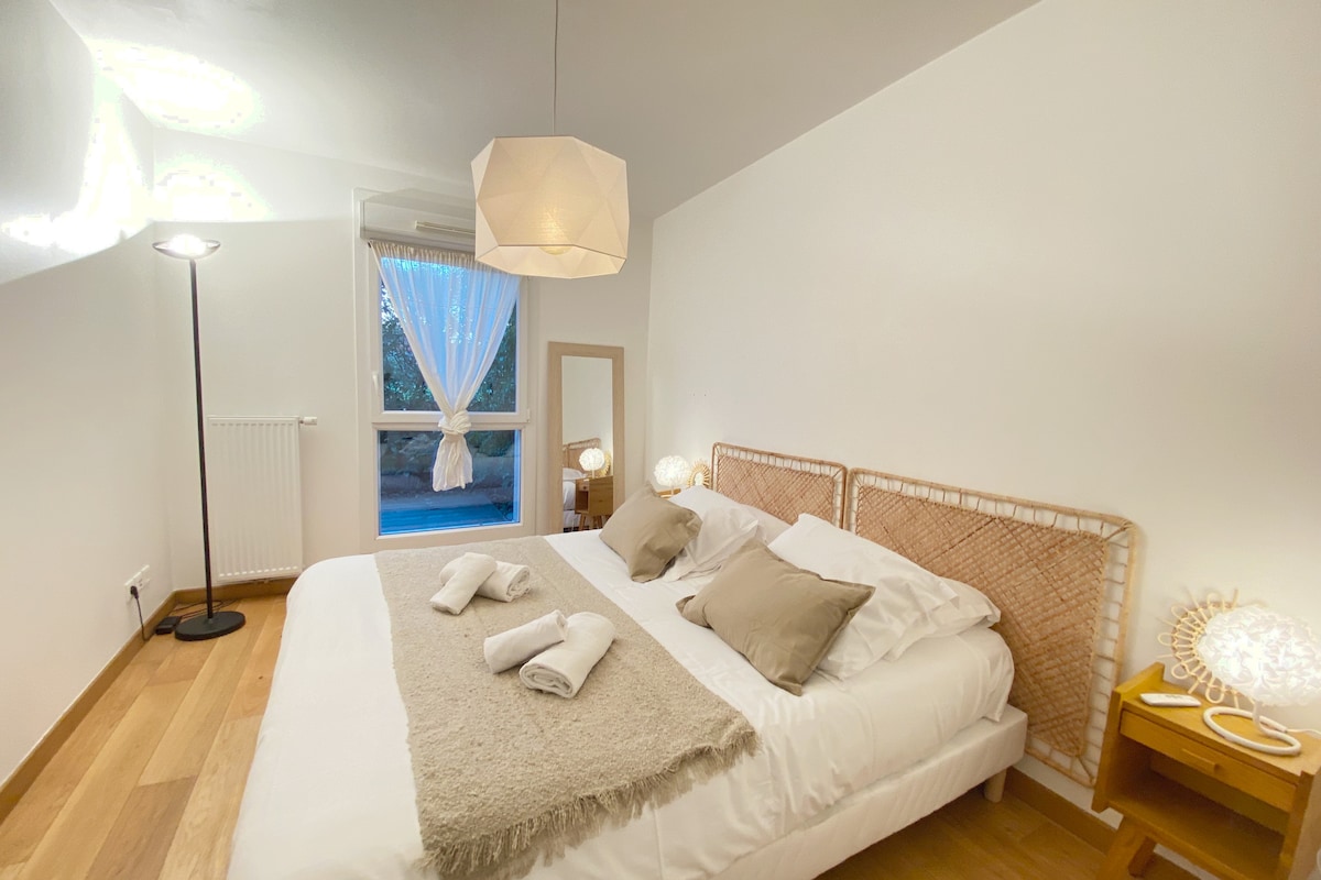 A welcoming bedroom features a spacious bed with neutral-colored linens and accent pillows. Soft lighting is provided by bedside lamps, and natural light filters through sheer curtains. A decorative mirror is positioned next to the window, creating an airy and bright ambiance.