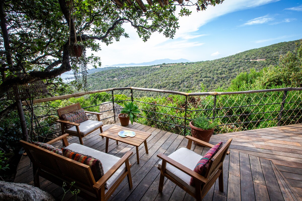 A wooden deck is surrounded by lush greenery, featuring a set of two armchairs and a matching coffee table. Views of rolling hills and a clear sky stretch into the distance, creating a peaceful outdoor seating area.
