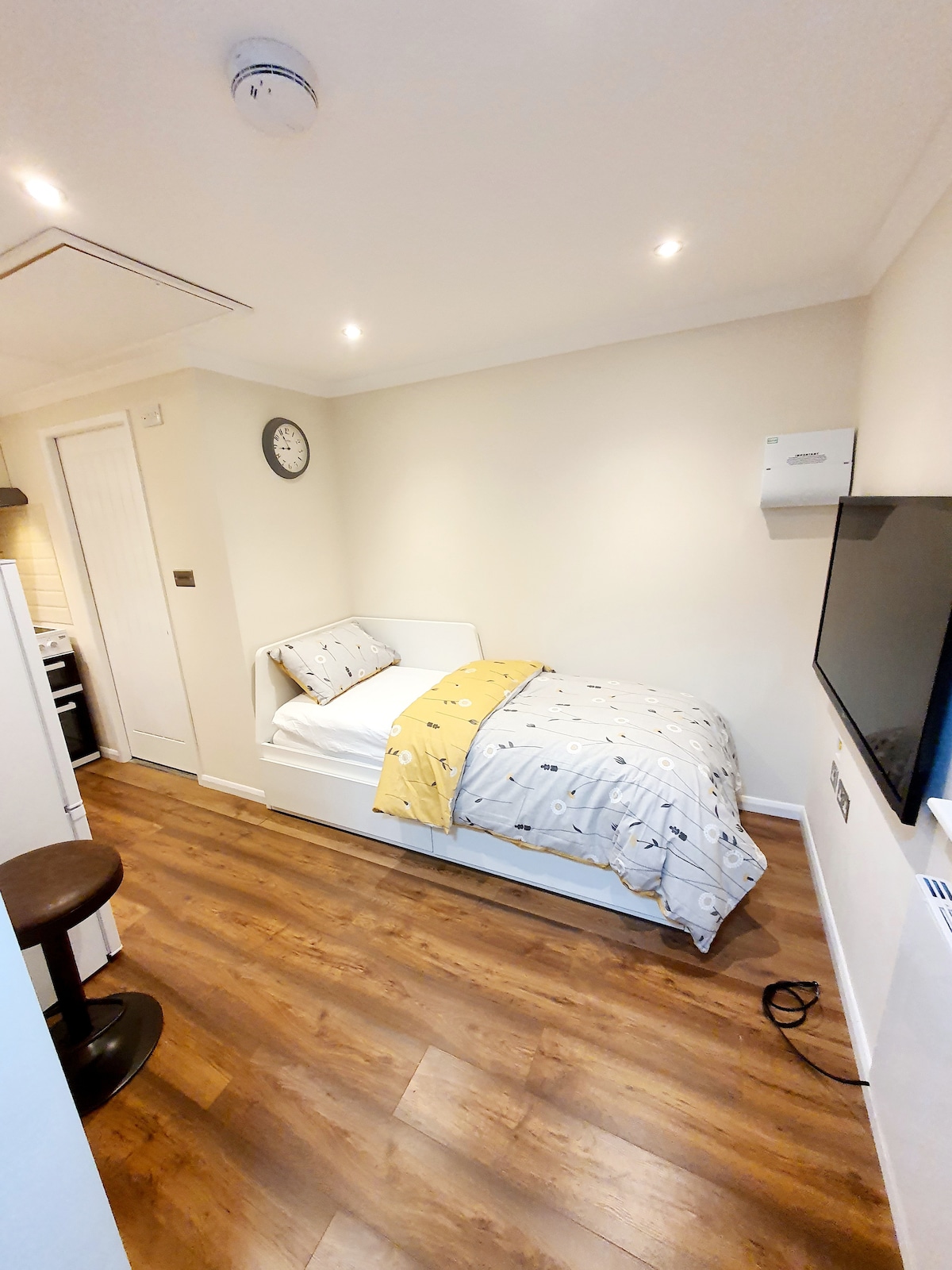 A single bed is positioned against a light-colored wall, adorned with patterned bedding. A wall clock is visible above the bed. A smart TV is mounted near the foot of the bed, with a wooden stool nearby and laminate flooring throughout.