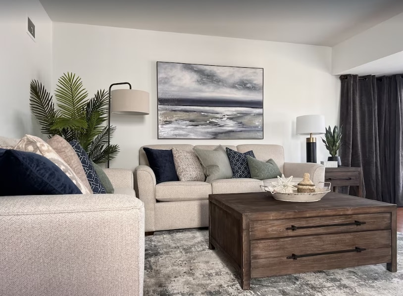 The living area is furnished with a light-colored sofa adorned with various decorative pillows, accompanied by a rustic wooden coffee table. A piece of abstract wall art is displayed above the sofa, and a floor lamp provides gentle illumination next to a leafy plant.