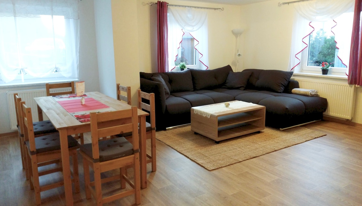 A spacious living area is furnished with a dark sectional sofa and a wooden coffee table, positioned on a natural fiber rug. A wooden dining table with six chairs is set nearby, and large windows allow natural light to enhance the open atmosphere.