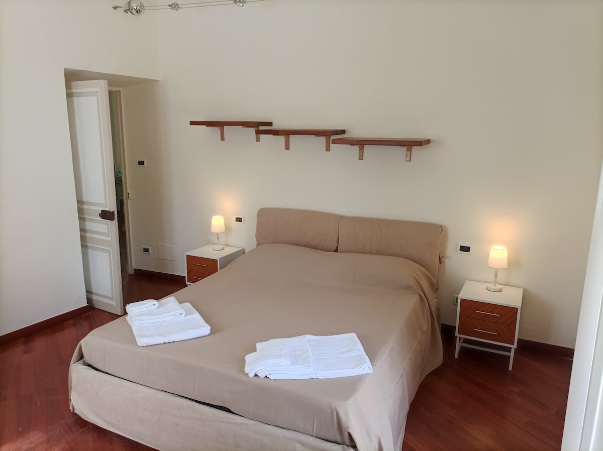 A spacious sleeping area features a neutral-toned bed dressed with fresh linens. Two bedside tables with soft lighting are positioned on either side. Decorative shelves line the wall above the bed, contributing to the room's overall simplicity.