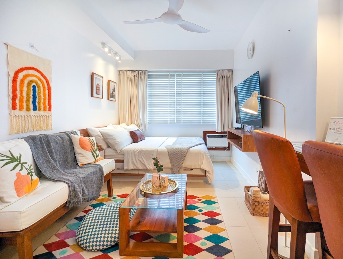 A bright and comfortable studio condo features a spacious seating area with a colorful rug, a coffee table, and modern entertainment setup. The natural light floods through the window draped in sheer curtains, illuminating the cozy sleeping area with two beds, tastefully paired with decorative elements.