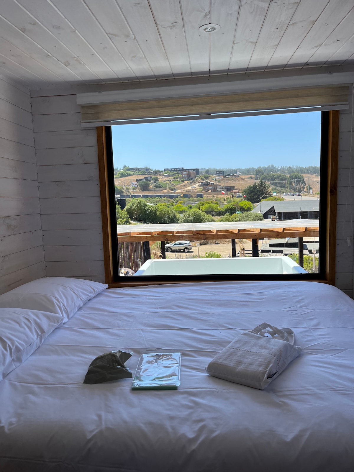 A bright sleeping area features a spacious bed with crisp white linens. A large window offers views of the scenic outdoor landscape, while a few items are placed neatly on the bed, including a towel and a small bag.