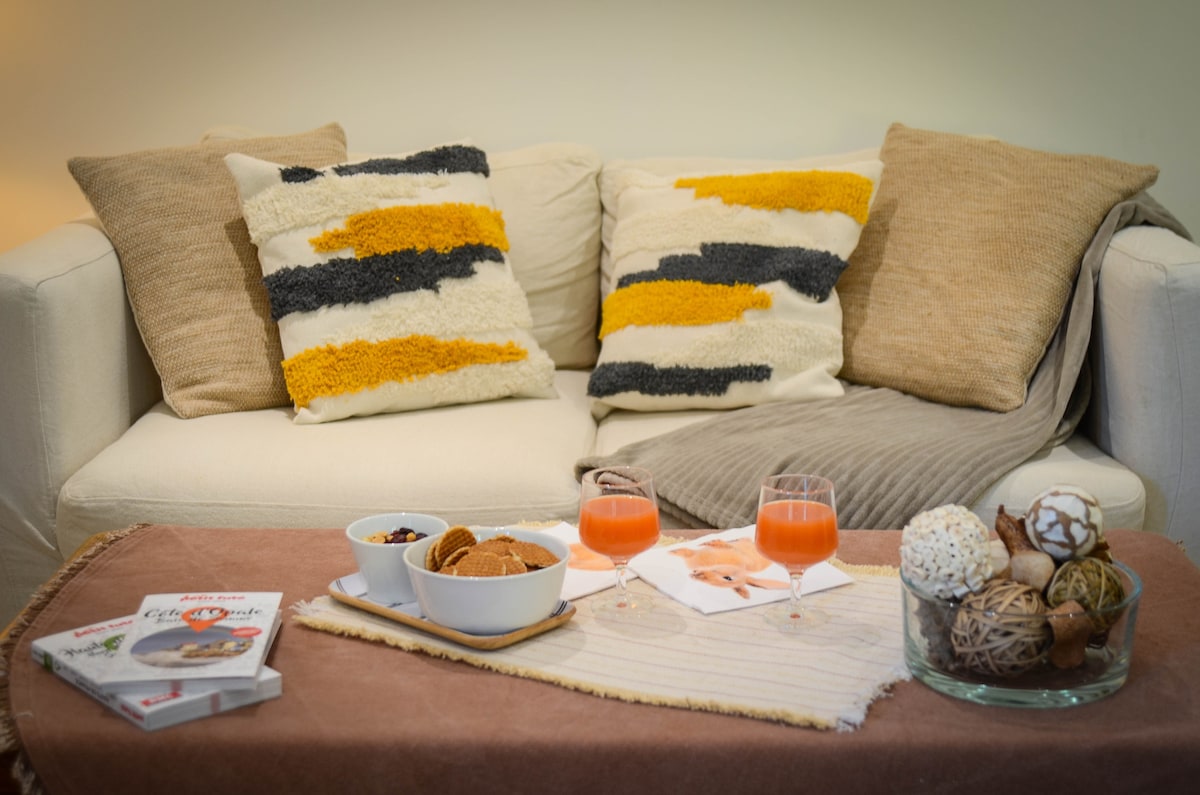 A neutral-toned sofa is adorned with textured cushions in yellow, gray, and beige. A low table features two glasses of juice, a bowl of snacks, and a magazine, alongside a decorative arrangement of natural materials and textured objects.