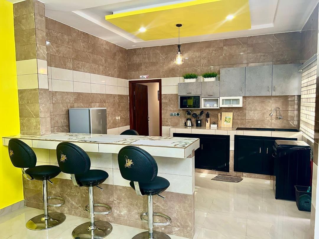 A modern kitchen offers a bar area with four high-backed stools beside a countertop. Neutral-toned cabinets contrast with bright yellow accents, while a refrigerator, microwave, and coffee maker are neatly arranged. Natural light filters through the window, enhancing the spacious feel.