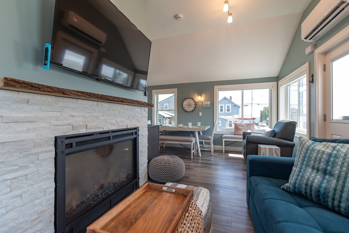 A modern living area features a stone fireplace and a large television mounted above. Comfortable seating is arranged around a coffee table, with a dining table visible nearby. Bright windows allow natural light and offer views of the surrounding area.