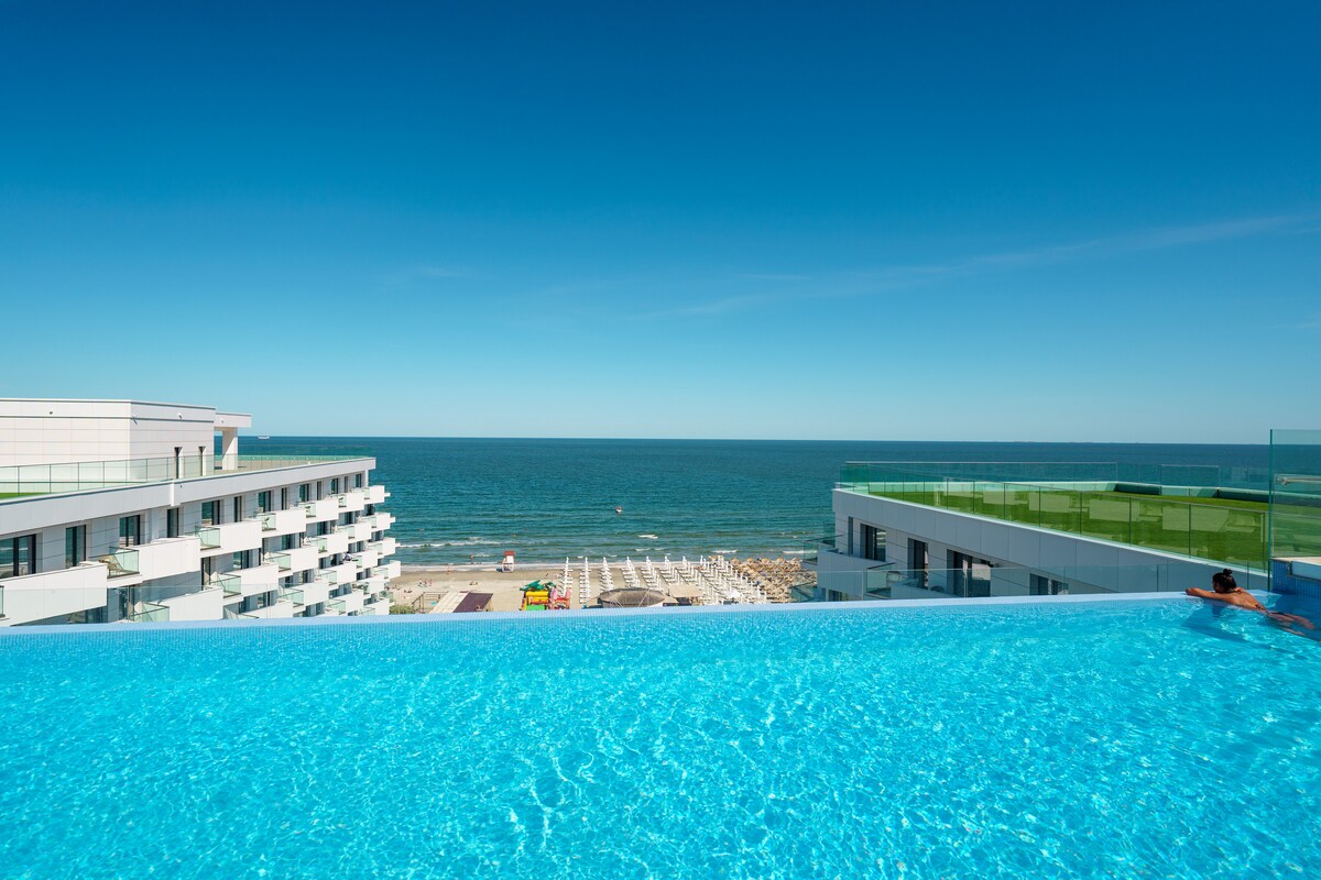 An inviting infinity pool is presented on the ninth floor, featuring clear blue water that extends towards the horizon. The ocean can be seen in the background, creating a serene backdrop against a bright blue sky. Nearby buildings outline the area, enhancing the view.