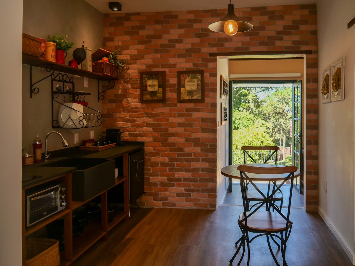 An open kitchen area features exposed brick walls and wooden shelving with decorative items. A dark sink is positioned next to appliances and utensils. A small dining table with two wooden chairs occupies the space, while large glass doors lead to an outdoor view of greenery.