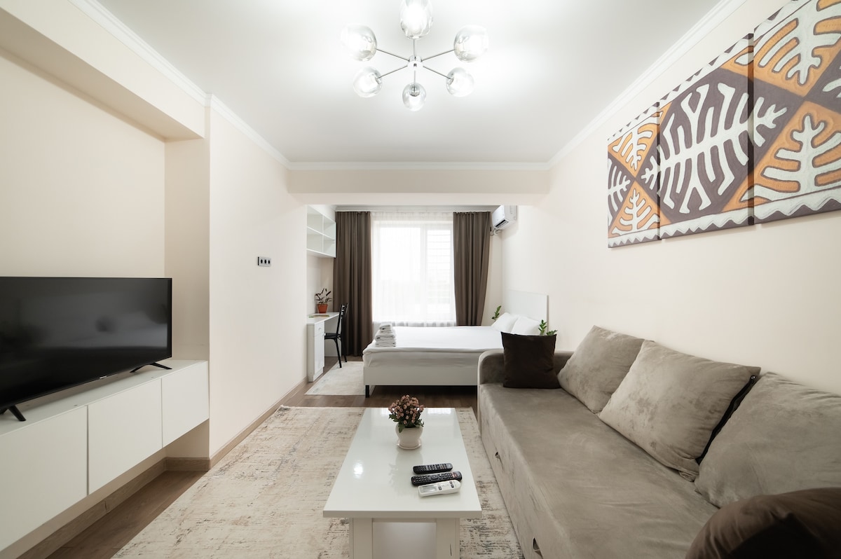 A light-filled living area features a comfortable sofa facing a flat-screen television. A coffee table is placed in front, while decorative elements such as a small plant add a touch of warmth. A spacious bed is visible in the background, complemented by soft curtains.